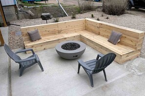 Pérgola de hormigón con zona de estar y banco de madera, dos sillas y un hogar circular.