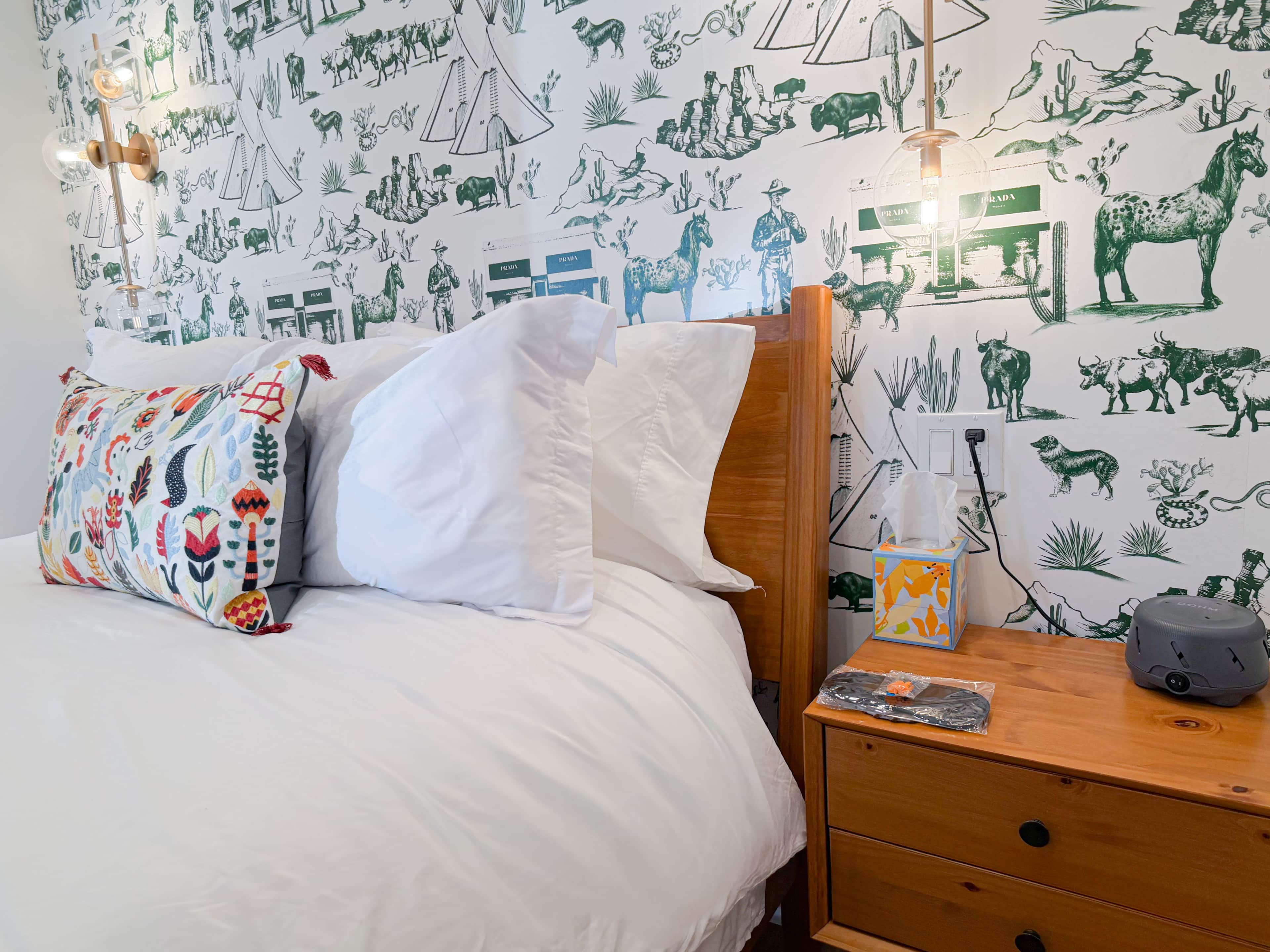 Un lit avec du linge de lit blanc, un coussin coloré, une table de chevet en bois avec une lampe, et un mur orné d’illustrations représentant la faune et la flore.