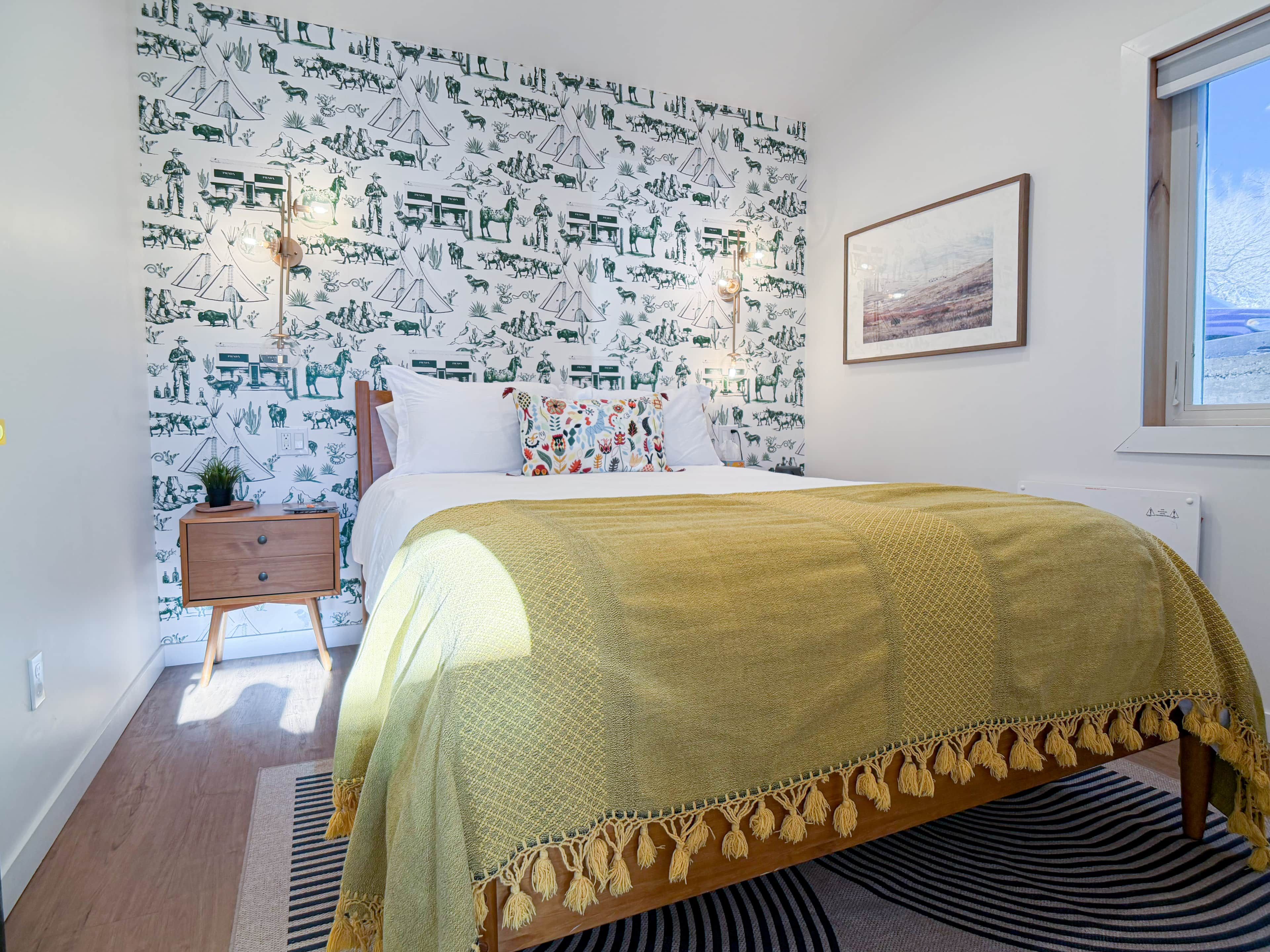 Une chambre avec un papier peint à motifs, un lit recouvert d’une couverture jaune, une table de chevet, une petite table avec une plante et un tableau encadré accroché au mur.