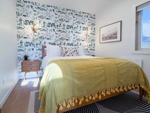 Um quarto com papel de parede estampado, uma cama com manta amarela, uma mesa de cabeceira, uma pequena mesa com uma planta e um quadro na parede.