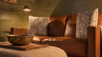 A brown leather sofa with floral cushions, a wooden bowl on a tray, and a lamp on the wall.