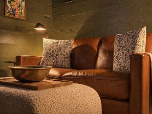 A brown leather sofa with floral cushions, a wooden bowl on a tray, and a lamp on the wall.