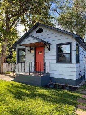 Une petite maison de plain-pied avec une porte rouge, un porche à l’avant et une cour clôturée.