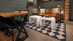 Ein Zimmer mit einem Holzespeisetisch, zwei gepolsterten Bänken, einem Ledersofa mit gemusterten Kissen und einem kleinen Beistelltisch, sowie einem karierten Teppich.