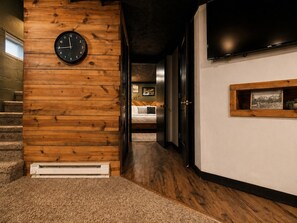 A hallway with wooden paneling, a clock, a bed with a checkered pillow, and a framed picture on the wall.