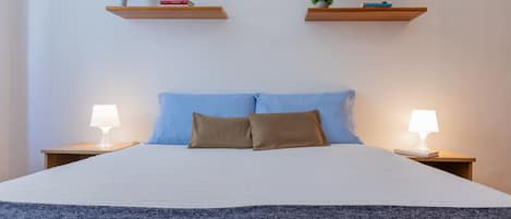A neatly made bed with a blue and brown pillow set, flanked by two bedside tables with lamps, and two wooden shelves above the bed.