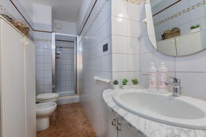 Un baño con lavamanos, inodoro y ducha blancos. Hay pequeñas macetas con plantas sobre el mostrador del lavamanos y un espejo encima.