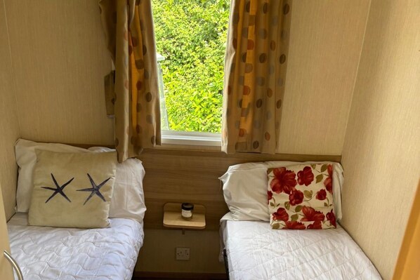 A compact RV bedroom with two beds, each with a distinct pillow design, a small table with a candle, and a window providing a view of greenery outside. - Seaside Lodge Haven in St Osyth (Clacton-on-Sea)