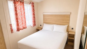 Une chambre avec un lit simple, une tête de lit en bois, du linge de lit blanc et une fenêtre agrémentée de rideaux à motifs floraux rouges.