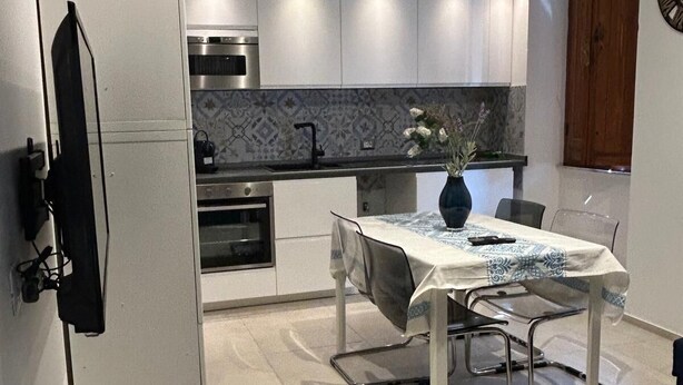 Una cocina moderna con armarios blancos, horno empotrado y encimera negra. Hay una mesa de comedor con sillas y un florero con flores.