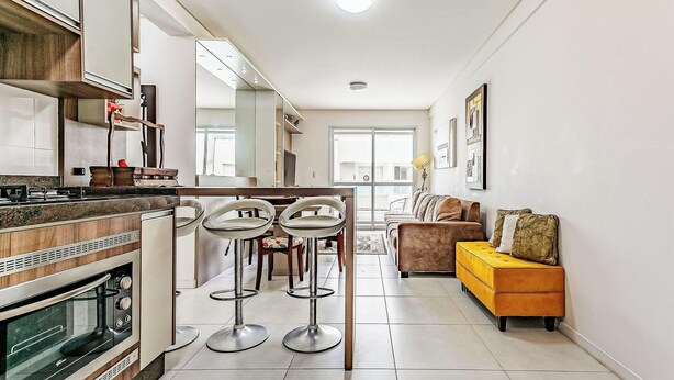 A modern kitchen with a built-in oven, a dining area with bar stools, a sofa, and a yellow ottoman.