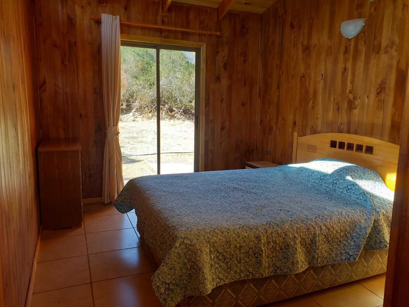 Una habitación de madera con una cama, un archivador pequeño y un ventanal corredero que da al exterior.