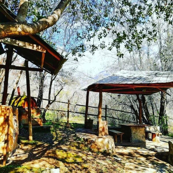 A rustic outdoor seating area with wooden tables and benches, surrounded by trees and a stone wall. - Cabañas Ocoa (Hijuelas)