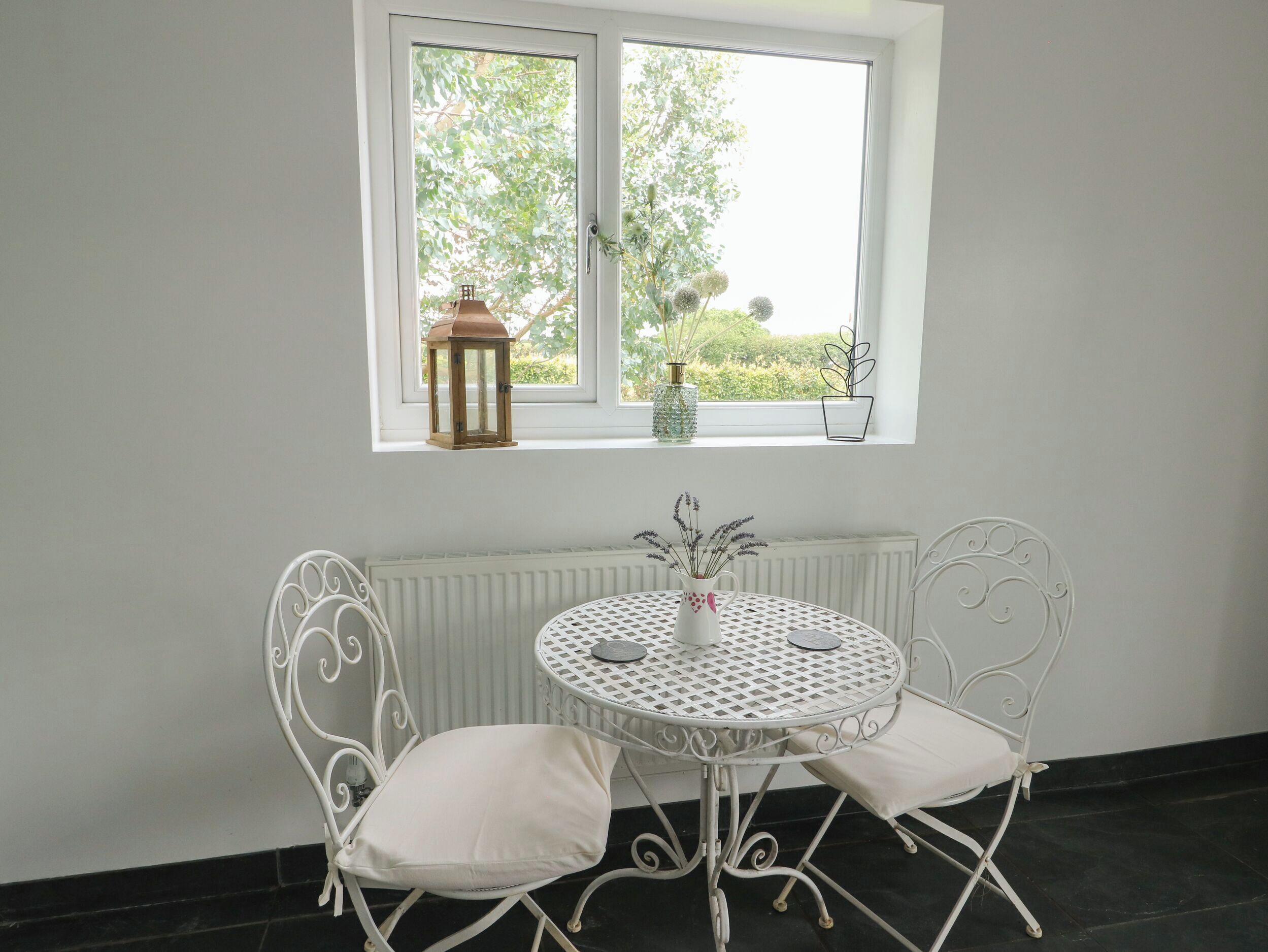 A white metal table with two chairs, a white radiator below, and a window with a view of greenery.
