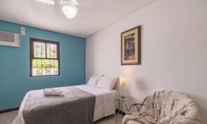 A bedroom with a bed, a window, a wall-mounted air conditioner, a framed picture, a bedside lamp, and a chair.