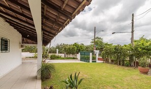 A covered patio area with a tiled floor, a grassy yard, and a white building with a brick wall. - Beautiful House 30m From Beach in Floripa Rtt1905 (Florianópolis)