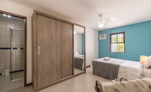 Une chambre avec un grand miroir, un lit recouvert d’une couverture grise, une table de chevet avec une lampe et une fenêtre donnant sur un paysage verdoyant.