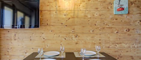 A dining table with four white plates and four wine glasses, set against a wooden wall with a TV and decorative items.