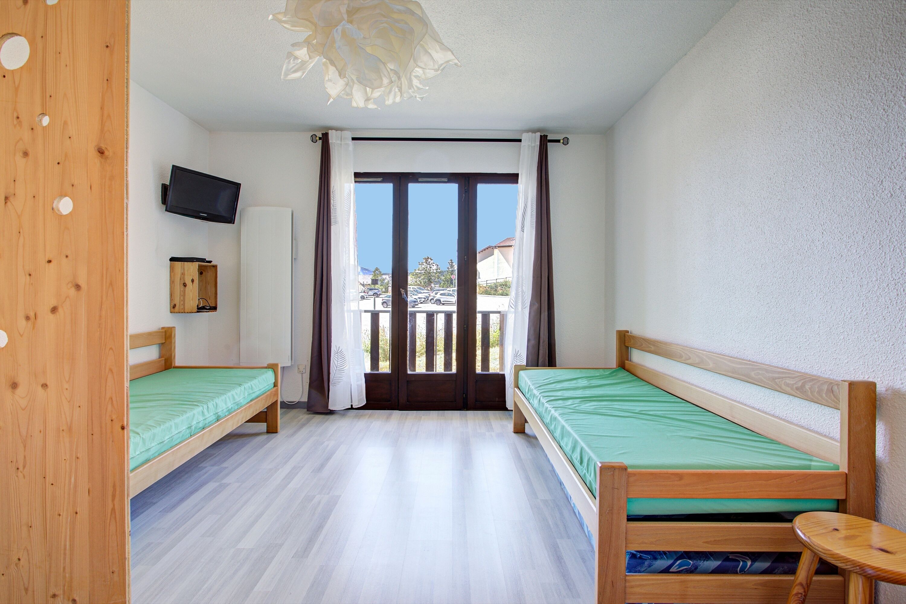 A room with two wooden beds, a television on the wall, and a balcony with curtains.