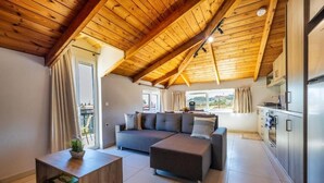A modern living room with a wooden ceiling, a grey sectional sofa, a wooden coffee table, and a kitchen area with stainless steel appliances. - Ionian Serenity House (Zakynthos)