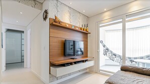 A modern living room with a TV on a wooden shelf, a sofa, and a large window with a view of a balcony. - Apto em prédio c/ piscina 350m da praia RTG208 (Florianópolis)