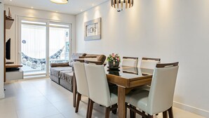 A modern dining area with a wooden table and white chairs, a sofa, and a large window with a view of a bridge. - Apto em prédio c/ piscina 350m da praia RTG208 (Florianópolis)