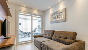 A living room with a brown sofa, a wooden TV stand, and a large window with a view of a staircase and outdoor decorations. - Apto em prédio c/ piscina 350m da praia RTG208 (Florianópolis)