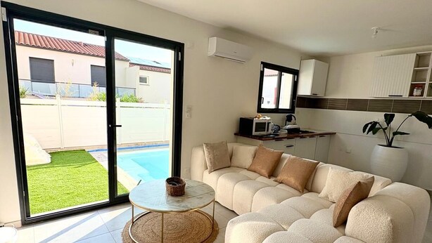 A modern living room with a sectional sofa, a small round table, and a view of a swimming pool through large glass doors. - Beautiful room in new villa with swimming pool (Perpignan)