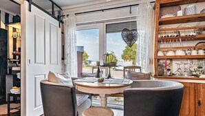 A dining area with a round table, two chairs, a small stool, and a wooden cabinet with dishes. - Apartment 'Kleine Oase Stuhr im Container E G' with Shared Terrace, Garden & Wi-Fi (Stuhr-Brinkum)