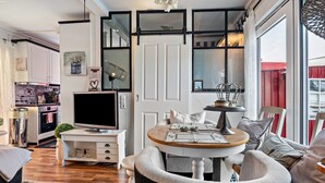 A modern kitchen with a flat-screen TV, a dining area with a round table, and a large window with a view. - Apartment 'Kleine Oase Stuhr im Container E G' with Shared Terrace, Garden & Wi-Fi (Stuhr-Brinkum)