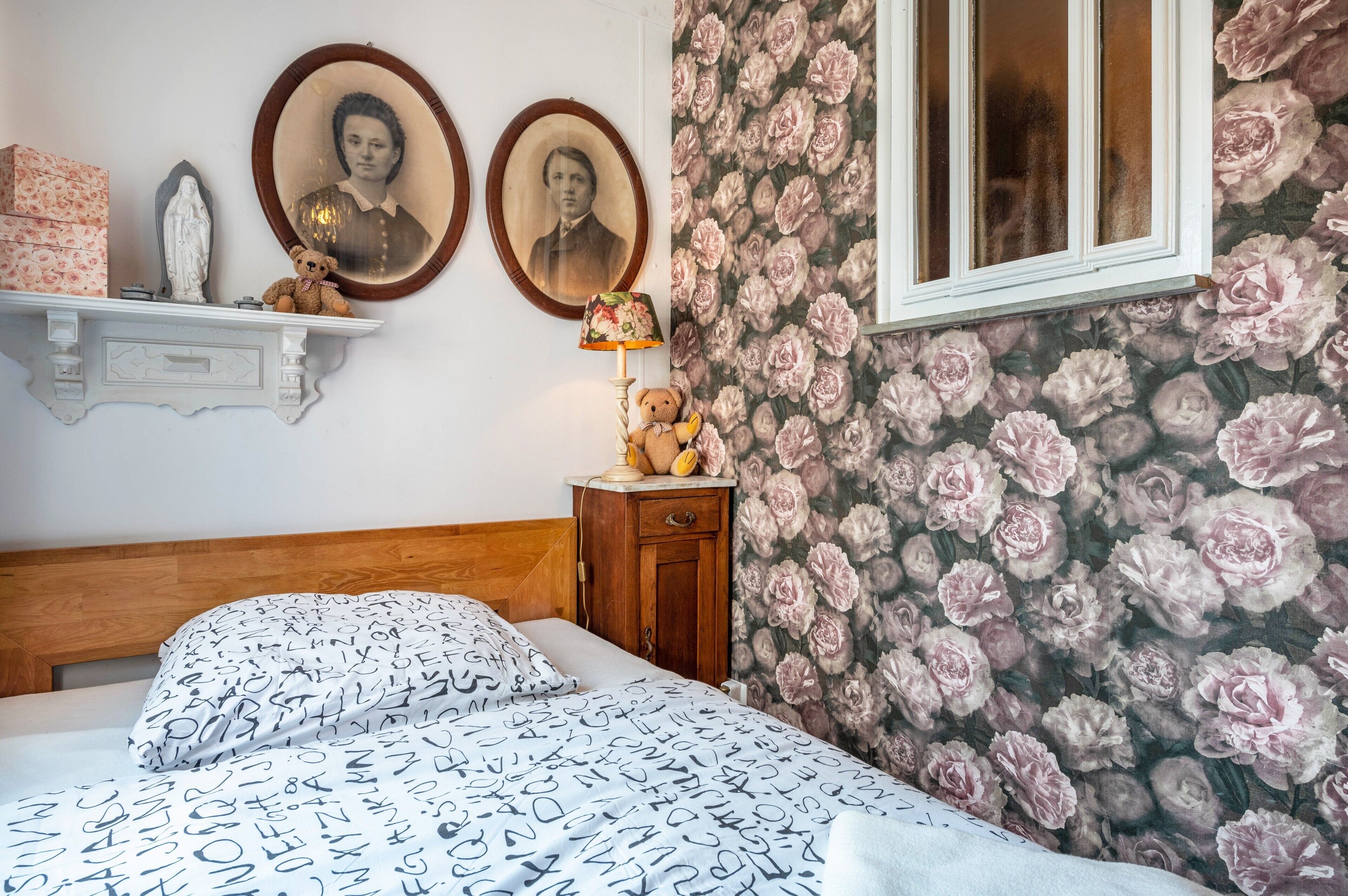 A bedroom with a bed, a dresser, a lamp, and floral wallpaper.