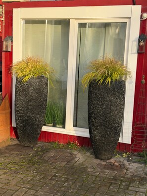 Deux grands bacs noirs, garnis de plantes, sont placés devant une fenêtre à cadre blanc surmontée d’une porte rouge.
