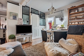 A modern living room with a flat-screen TV, a dining area with a table and chairs, a wicker chair, a chandelier, and a shelf with decorative items. - Apartment 'Kleine Oase Stuhr im Container E G' with Shared Terrace, Garden & Wi-Fi (Stuhr-Brinkum)