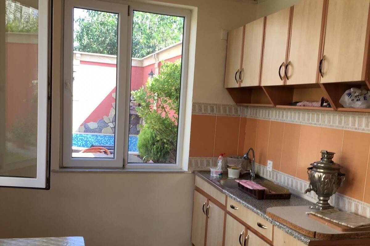 A kitchen with wooden cabinets, a window with a view of greenery, a tiled floor, and a countertop with a kettle and a cup.