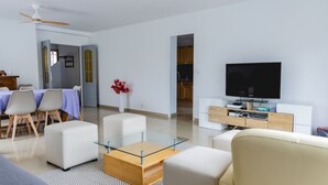 Ein modernes Wohnzimmer mit einem Glastisch, weißen Sofas, einem Flachbildfernseher und einem Essbereich mit Stühlen und Tisch.