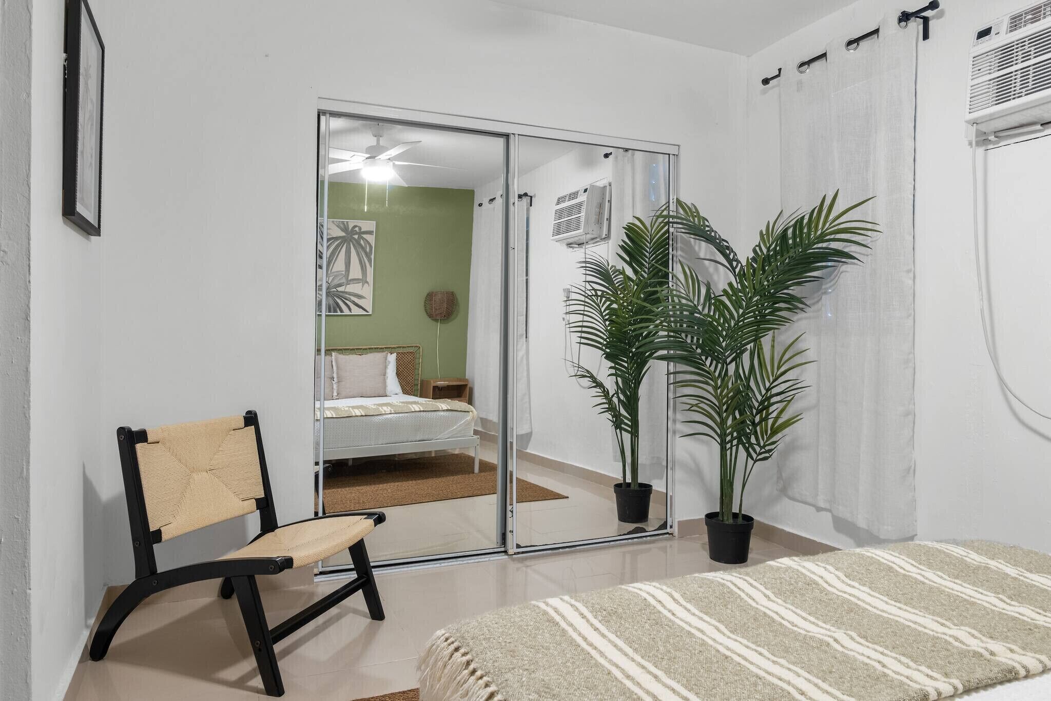 Un dormitorio con una cama, una silla, un espejo grande y dos plantas en macetas.