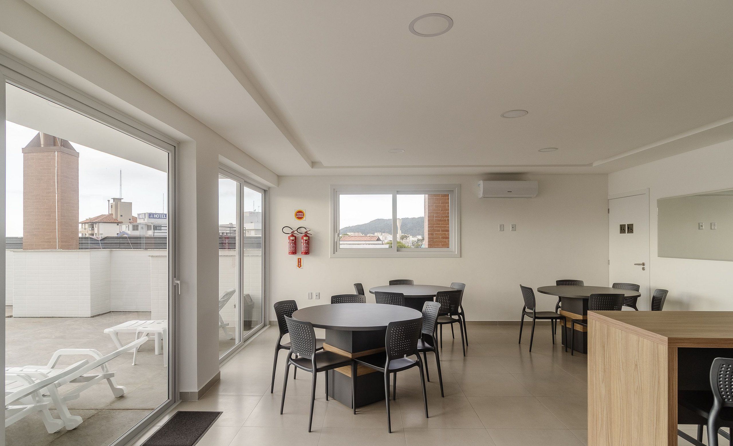Una habitación de hotel moderna con un amplio ventanal corredero que da a un balcón, una mesa de comedor redonda con sillas y una pequeña zona de kitchenette.