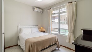 Ein Schlafzimmer mit Bett, Deckenventilator, Klimaanlage, Nachttisch mit Lampe und einem Fenster mit Vorhängen.