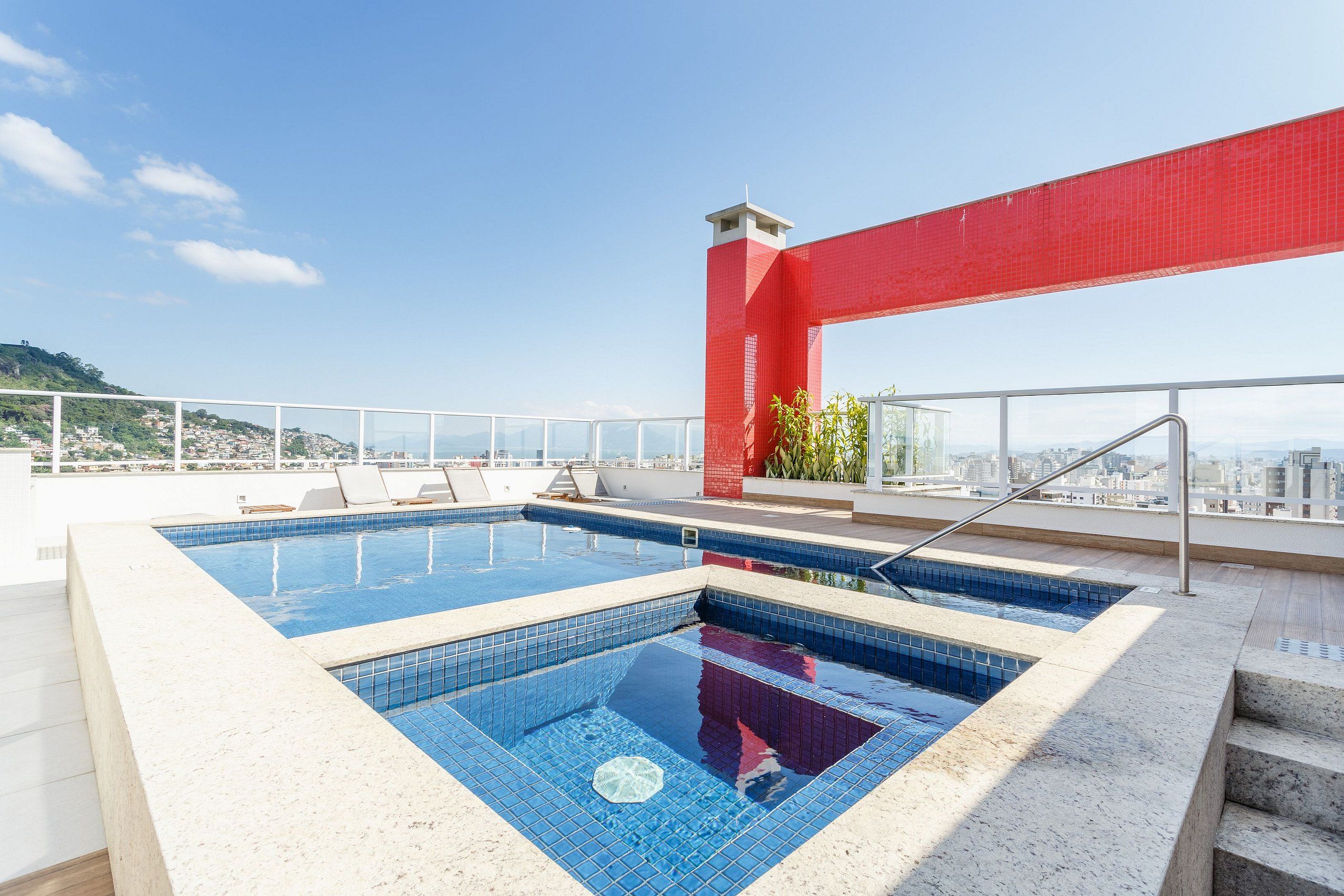 A rooftop pool with clear blue water, a red structural element, and a city view in the background.