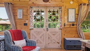 A cozy cabin interior with a wooden ceiling, a plaid armchair with a red throw, a cowhide rug, and a rustic door with a welcome sign. - The Nest-A Cozy Rustic Escape for Two (Fredericksburg)