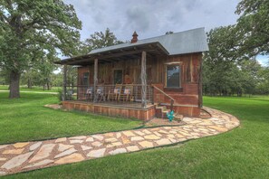 A rustic cabin with a porch, stone pathway, and a fenced area with chairs. - The Roost - Charming Hill Country Hideaway (Fredericksburg)