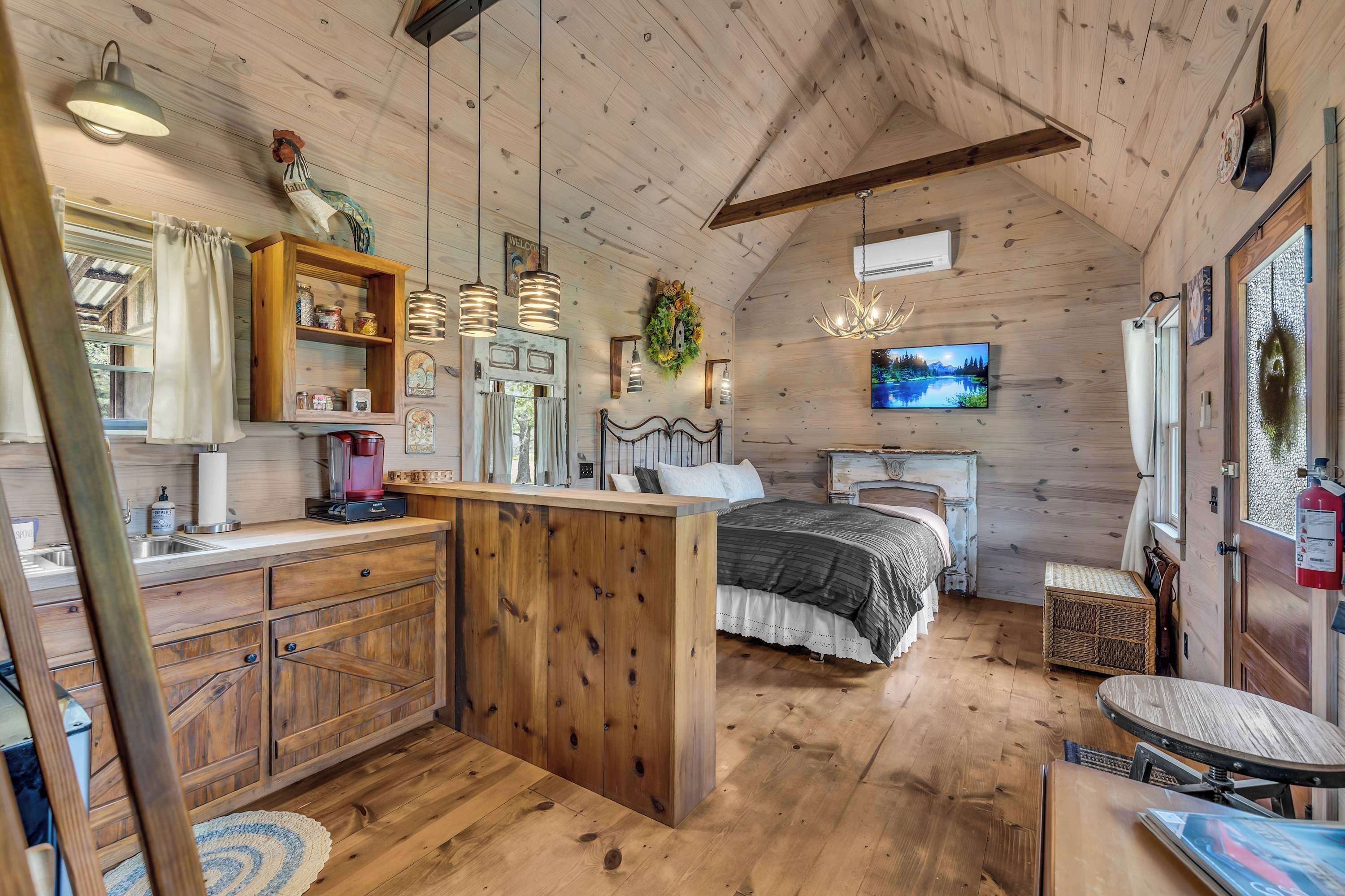 A cozy cabin interior with a wooden ceiling, a kitchen area with a wooden island, a bed with a view, and a mounted TV.