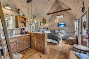 A cozy cabin interior with a wooden ceiling, a kitchen area with a wooden island, a bed with a view, and a mounted TV.