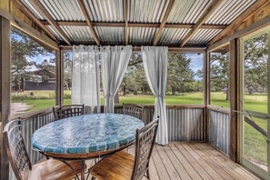 A covered outdoor seating area with a round table and chairs, large windows, and a view of a grassy landscape.