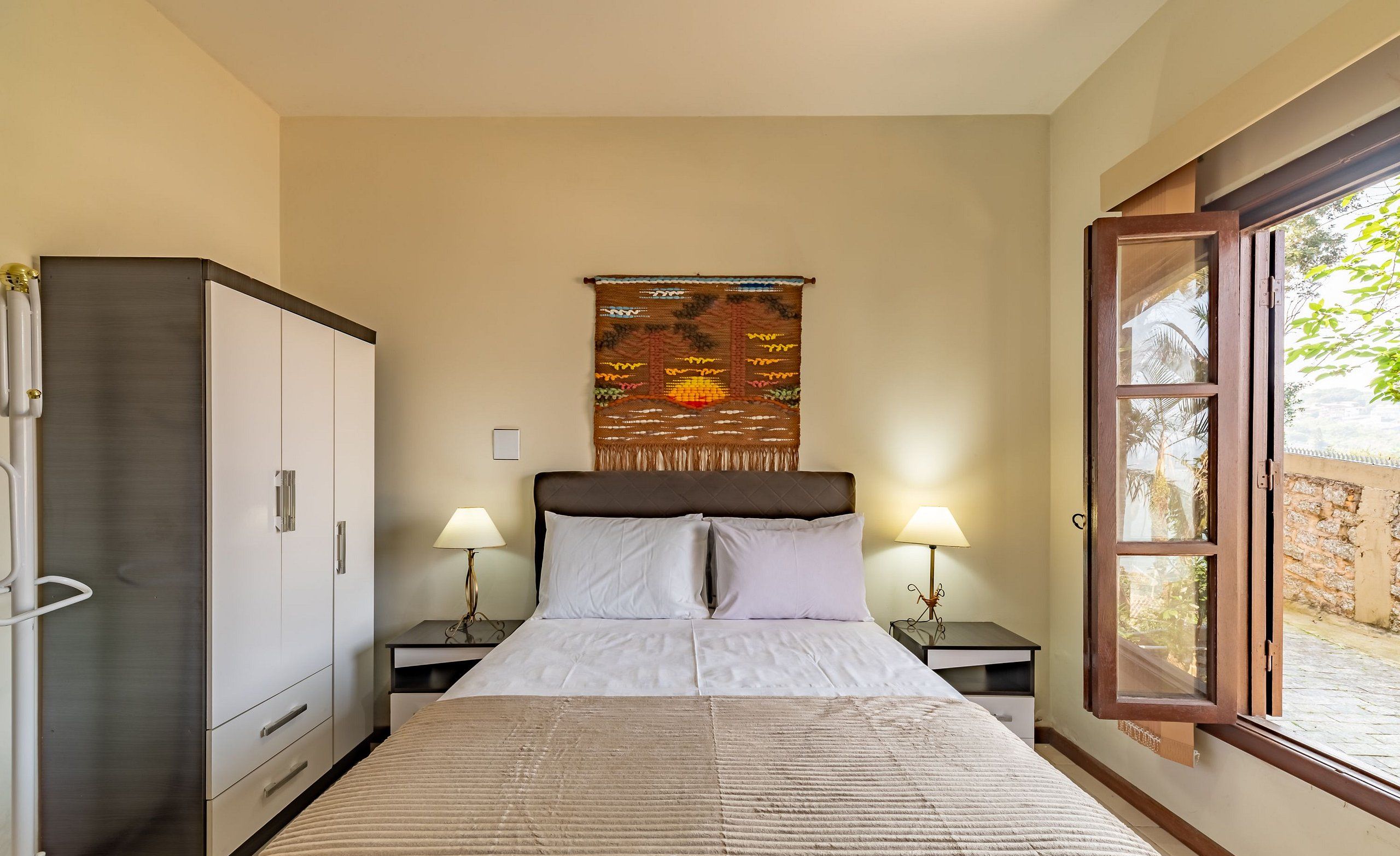 A bedroom with a bed, nightstands, a wardrobe, a tapestry, and a window with a view.