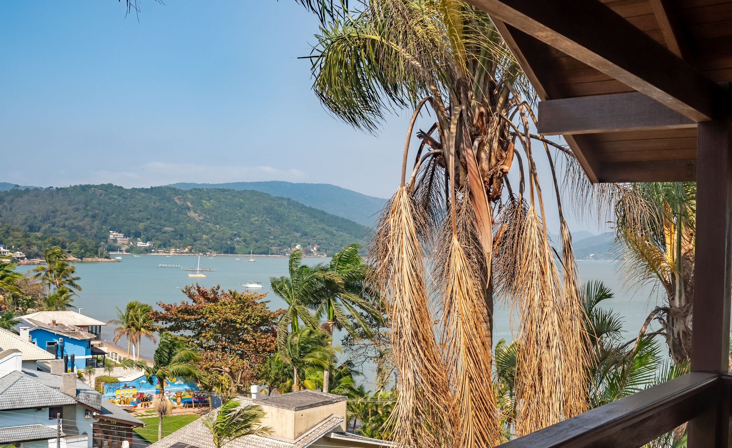 A coastal view with a wooden balcony overlooking a beach, palm trees, and a distant mountain range.