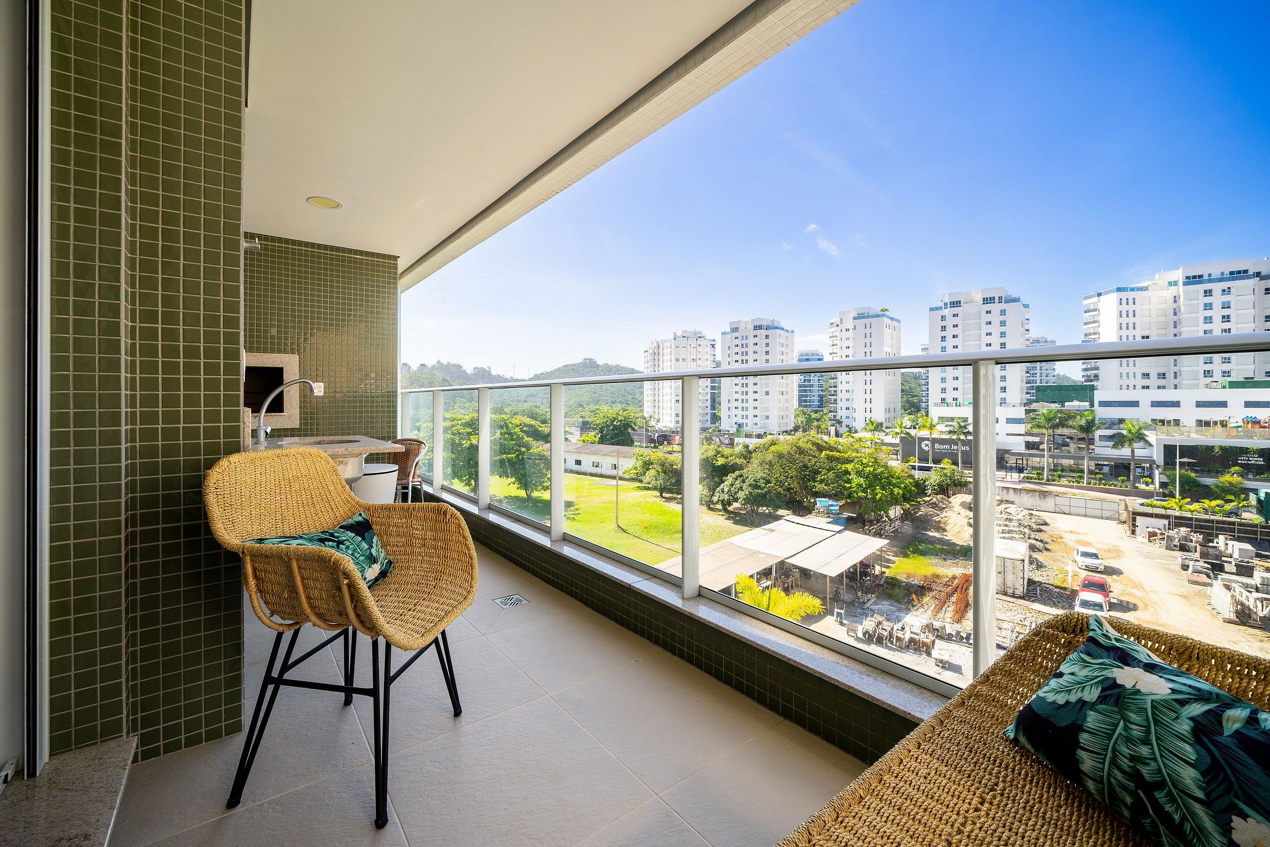 Un balcón con una silla de mimbre y un sofá, una pared con azulejos y vistas a edificios y zonas verdes.