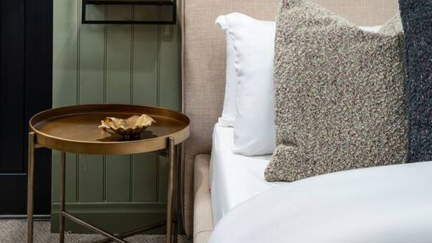A hotel room with a bed, a bedside table with a lamp, a shelf with a mirror, and a door.