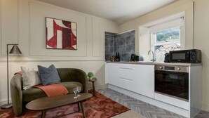A compact kitchen with white cabinets, a built-in oven, and a microwave. There's a small table with a glass, a sofa with cushions, and a painting on the wall.