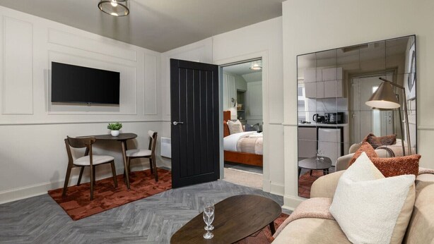 Ett modernt hotellrum med en platt-TV, en matplats med bord och stolar, en soffa med kuddar och ett kök som syns genom en glasdörr.